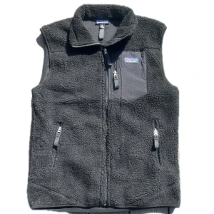 Svart fleeceväst från Patagonia - Mysig svart fleeceväst från Patagonia med hög krage och tre praktiska fickor med dragkedja, varav en bröstficka. Västen har en enkel, stilren design och är tillverkad i mjuk fleece som håller dig varm. Perfekt för lager-på-lager under kyliga dagar.