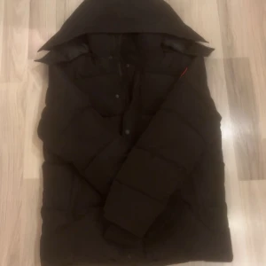Svart pufferjacka från Canada Goose - Svart pufferjacka från Canada Goose med klassisk logga på ärmen. Jackan har stor huva, långa ärmar och knappar framtill. Insidan är fodrad med glansigt material och jackan är perfekt för kalla dagar. Snygg och stilren design som håller dig varm. Helt ny var tyvärr för stor 