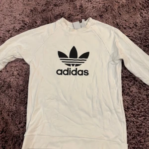 Vit Adidas sweatshirt med logga - Vit långärmad sweatshirt från Adidas med stor svart trefoil-logga och text på bröstet. Tröjan har rund hals och är tillverkad i mjuk bomull, perfekt för chill dagar eller när du vill ha en clean streetstyle-look.