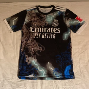 Real Madrid Bellingham draktröja - Säljer en Real Madrid fotbollströja med Bellingham och nummer 5 på ryggen. Svart bas med detaljerade drakmotiv i grått och blått, vita ränder på axlarna och tryck från Emirates. Tillverkad i lätt polyester, kortärmad och med rund hals.
