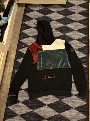 Färgglad hoodie från Vichy med huva - Unik hoodie från Vichy med svart bas och färgblock i vinrött, mörkgrönt och vitt på framsidan. Tröjan har broderad arabisk text, huva med snörning och en stor magficka. Materialet är mjukt och känns som bomull och sammet. Perfekt för dig som gillar streetwear med personlig touch.