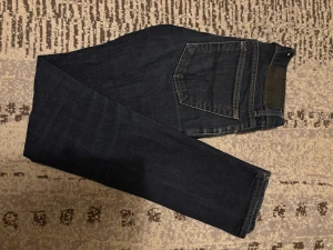 Mörkblå Tiger of Sweden jeans 27/32 - Säljer ett par mörkblå jeans från Tiger of Sweden i modellen Base. Klassisk femficksdesign, raka ben och snygga kontrastsömmar. Jeansen är tillverkade i slitstarkt denim och har en stilren look som passar till det mesta.