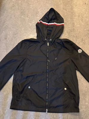 Svart vindjacka från Moncler med huva - Svart vindjacka från Moncler med huva och snörning, dragkedja framtill och två fickor med tryckknappar. Jackan har en vit och röd rand på huvan och Moncler-logga på ärmen. Materialet är lätt och syntetiskt, perfekt för blåsiga dagar.