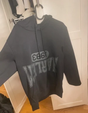 Svart hoodie från H&M med tryck - Svart hoodie från H&M i regular fit med stort tryck på framsidan där det står 'HARLEM 1983' i grått. Tröjan har huva med snörning och långa ärmar. Perfekt för dig som gillar streetstyle och vill ha en chill look.