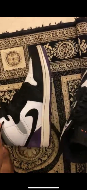 Nike Air Jordan 1 svart/vit/lila - Säljer ett par Nike Air Jordan 1 med klassisk high-top siluett. Skorna har svart och vit ovandel med lila detaljer på sulan. Ikonisk swoosh på sidan och snörning framtill. Perfekta för dig som vill sticka ut med en streetwear vibe.