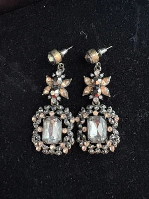 Glittriga örhängen med stenar - Säljer ett par glittriga örhängen i silverfärg med stora rektangulära stenar och små runda kristaller i beige och silver. Örhängena har en blomformad detalj upptill och är riktigt statement med sin gnistrande look. Perfekta för dig som gillar att sticka ut.