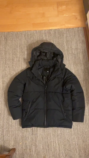 Svart pufferjacka med huva - Svart pufferjacka i klassisk modell med huva och dragkedja framtill. Jackan har en enkel, stilren look med två sidofickor och breda kanaler. Perfekt för kalla dagar och riktigt skön att bära. den är i storlek 150 men sitter ungefär 165-175