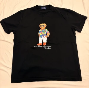 Polo Ralph Lauren – Polo Bear T-shirt (L) NY - Säljer en helt ny, oanvänd Polo Bear T-shirt från Polo Ralph Lauren i storlek L. Svart modell med ikoniska “Polo Bear”-trycket i färg och texten Polo Bear by Ralph Lauren framtill. Mjuk bomullskvalitet, perfekt passform och aldrig använd – endast uppackad för foto.  ✔ Storlek: L ✔ Skick: Helt ny / oanvänd ✔ Färg: Svart