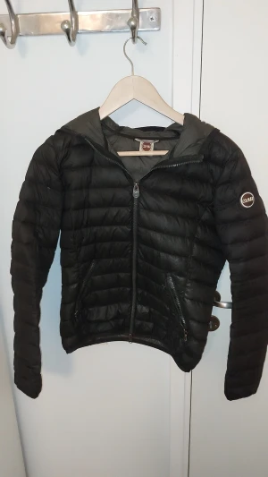 Svart dunjacka från Colmar med huva - Strl 42 skulle säga att passar barn 164. Svart dunjacka från Colmar med huva och quiltad design. Jackan har dragkedja framtill, två sidofickor med dragkedja och ett Colmar-märke på ärmen. Lätt och smidig modell som passar perfekt för kyliga dagar. Snygg, stilren och enkel att matcha. Kan gå ner I pris. Skick 6/10 se bilder den har ett hål som är lagat märks inget.