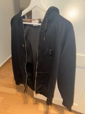 Svart Burberry hoodie med dragkedja - Svart hoodie från Burberry i storlek XS då den tvättats o blivit mindre .med klassisk rutig insida på huvan. Tröjan har broderad Burberry-logga på bröstet, snörning i huvan och praktiska fickor framtill. Materialet är mjukt och känns lyxigt, perfekt för dig som gillar stilrena streetwear-plagg.