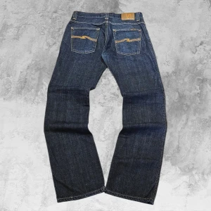 Rare Premium Nudie Dark Indigo Jeans - Dessa Nudie Jeans har en snygg mörk indigotvätt och det klassiska Nudie-våg-stygnet på bakfickorna. Rakt och bekvämt passform med den välkända Nudie-kvaliteten. De orange kontrastsömmarna ger en riktigt fin detalj som sticker ut.  Mått: Total längd: 103 cm Midja: 40 cm Benöppning: 19 cm  Ett par riktigt bra och hållbara kvalitetsjeans från ett märke som förtjänar en plats i garderoben. Passa på innan någon annan hinner :)