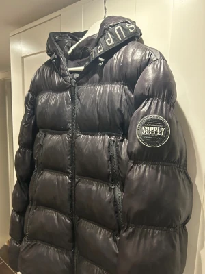 Svart pufferjacka Supply & Demand - Svart pufferjacka från Supply & Demand med glansig finish och tydlig logga på ärmen. Jackan har huva med broderad text, dragkedjor framtill och två stora fickor. Perfekt för kalla dagar när du vill hålla stilen.