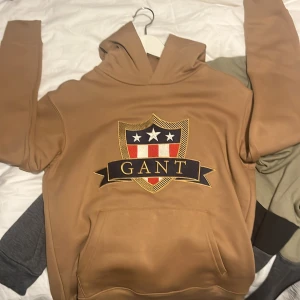 Beige hoodie från GANT med emblem pris kan diskuteras (15 år ) - Säljer en snygg beige hoodie från GANT med stort broderat emblem på bröstet i blått, rött, vitt och guld. Tröjan har huva och känguruficka framtill. Perfekt för dig som gillar klassisk och sportig stil.