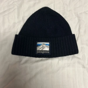 Svart ribbstickad mössa Patagonia - Svart ribbstickad mössa från Patagonia med uppvikt kant och snyggt broderat logomärke framtill med bergmotiv i grått, blått och vitt. Klassisk form som sitter skönt på huvudet 10/10 skick knappt använts.