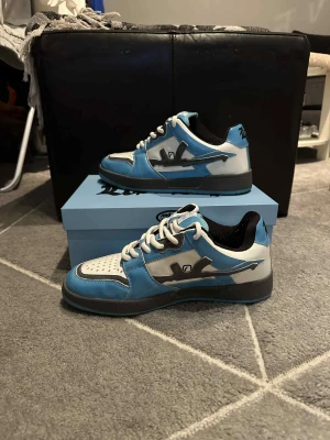 Lost Boys Electric Sky sneakers blå/vit - Lost Boys Electric Sky sneakers i blått och vitt med svarta detaljer och grå inslag. Låg modell med snörning, perforerad tåbox med små kors, och svart yttersula. Märkets logga på sidan och 'Lost Boys' broderat på plösen. Snygg streetstil för dig som gillar unika sneakers.