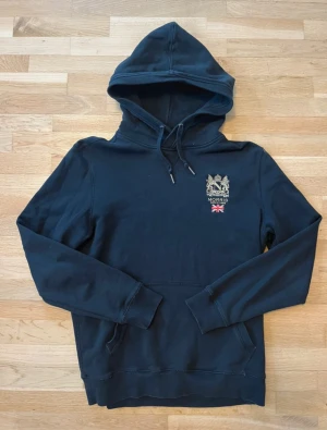 Svart tröja med luva från Morris med broderi - Svart hoodie från Morris med snyggt broderat emblem och brittisk flagga på bröstet. Tröjan har huva med snörning, känguruficka och långa ärmar. Perfekt för dig som gillar klassisk stil med en twist.
