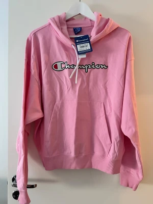Rosa Champion hoodie med logga - Säljer en rosa hoodie från Champion med stor logga framtill. Tröjan har huva med snörning, magficka och ribbade muddar vid ärmslut och nederkant. Perfekt för dig som gillar sportig och avslappnad stil.