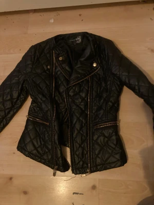 Svart quiltad skinnjacka Attentif - Snygg svart quiltad skinnjacka från Attentif med guldiga dragkedjor och knappar. Jackan har figurnära passform, långa ärmar och flera fickor med dragkedja. Perfekt för dig som gillar edgy stil och detaljer som sticker ut.