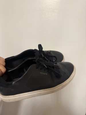 Svarta Arigato sneakers  - Snygga svarta sneakers från Arigato med stilren design och vit platt sula. Skorna har rund tå, svarta snören och är tillverkade i skinn med diskret guldfärgad logga på sidan. Perfekta för dig som gillar minimalistisk stil och vill ha ett par schyssta sneakers.