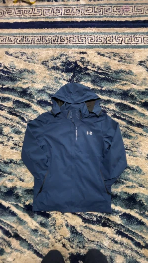 Blå vindjacka från Under Armour - Snygg blå vindjacka från Under Armour med huva och dragkedja.Jackan har en stilren look med logga på bröstet och diskreta detaljer. Perfekt för dig som vill ha något som står emot vind och väta.(Jackan är vattentät)