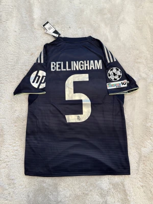 Real Madrid Bellingham #5 fotbollströja - Snygg mörkblå Real Madrid fotbollströja med Bellingham och nummer 5 på ryggen. Adidas-logga, Champions League-märke och HP-sponsor på ärmarna. Guldiga detaljer och broderade klubbmärken. Tillverkad i lätt och ventilerande material.