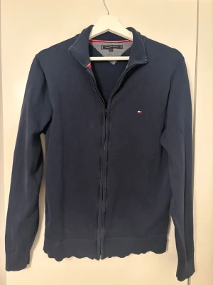 Marinblå kofta från Tommy Hilfiger - Snygg marinblå kofta från Tommy Hilfiger med hel dragkedja framtill och diskret logga på bröstet. Tröjan har långärmad design och ribbade muddar. Materialet är mjuk bomull och insidan har ett mönstrat tyg vid kragen. Perfekt för dig som gillar klassisk stil.