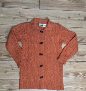 Rostorange kofta från Aran Crafts - Säljer en sjukt snygg rostorange kofta från Aran Crafts, tillverkad i 100% merinoull. Koftan har ett grovt stickat mönster, stora svarta knappar och bred krage. Perfekt för dig som gillar klassisk stil med en twist. Made in Ireland.