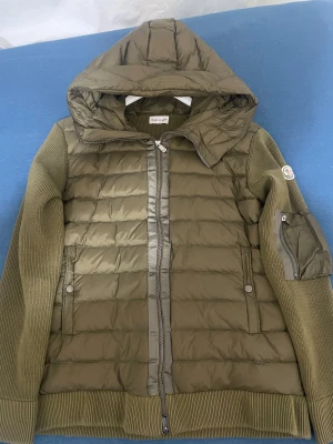 Olivgrön Moncler cardigan  - Snygg olivgrön pufferjacka från Moncler med stickade ärmar och detaljerad ficka med dragkedja på armen. Jackan har quiltad front, två sidofickor med tryckknappar och en stor huva. Perfekt för dig som vill ha en cool och funktionell look.