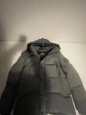 Grå vinterjacka från Canada Goose - Säljer en grå vinterjacka från Canada Goose med huva och stora fickor. Jackan har en snygg patch på ärmen, dragkedja och knappar framtill. Perfekt för kalla dagar och har en clean, puffad look som är riktigt chill.Om ni har några frågor eller om ni vill se mer bilder tveka inte hör av er😀och om ni undrar om den är äkta så är den de 😀