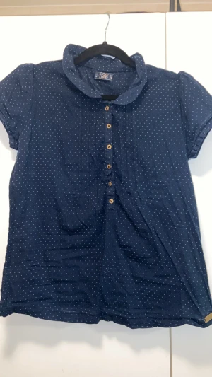 Mörkblå prickig henley från P&B - Snygg mörkblå henley-skjorta från P&B med vita prickar och träfärgade knappar framtill. Skjortan har korta ärmar, mjuk krage och är gjord i lätt bomullstyg. Perfekt för dig som gillar en chill och stilren look.