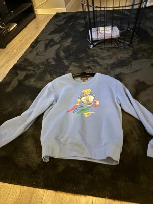 Blå sweatshirt från Polo Ralph Lauren - Säljer en ljusblå sweatshirt från Polo Ralph Lauren med ett stort tryck av en björn som målar färgglada penseldrag på framsidan. Tröjan har rund hals, långa ärmar och är gjord i mjuk bomull. Perfekt för dig som gillar streetwear och coola prints.