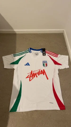 Italien x Stüssy t-shirt - Snygg vit t-shirt från Adidas i samarbete med Stüssy. Tröjan har gröna och röda detaljer på sidorna, blå krage, broderat Italienskt landslagsmärke och Adidas-logga samt Stüssy-tryck i rött framtill. Slim fit och tillverkad i polyester.