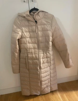 Beige lång pufferjacka Chiara Forthi XS - Säljer en beige lång pufferjacka från Chiara Forthi i storlek XS. Jackan har huva, quiltad design och dragkedja hela vägen fram. Perfekt för kalla dagar och har en snygg, stilren look med figurnära passform.