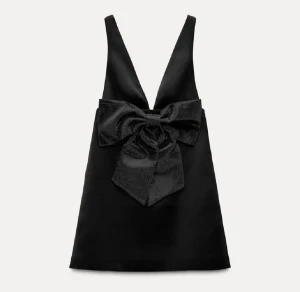 Nyår klänning - Supersnygg svart kortklänning med djup v-ringning och en stor glittrig rosett framtill. Klänningen är ärmlös och har en stilren, festlig look med lätt A-linjeform. Perfekt för dig som vill sticka ut och älskar statement-detaljer.