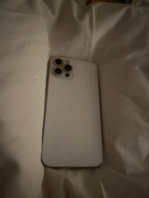 iPhone 12 Pro - Säljer en iPhone 12 Pro i silver. Mobilen har tre kameralinser och LiDAR-sensor. Baksidan är sprucken med tydliga sprickor, men kamerorna ser hela ut. Skärmen ser ut att vara i bra skick utan synliga sprickor. Perfekt för dig som vill ha en kraftfull iPhone till ett bra pris.
