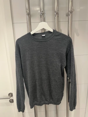 Mörkgrå stickad tröja i merinoull  - En enkel och stilren mörkgrå stickad tröja med rund hals och långa ärmar. Tröjan har ribbade muddar vid hals, ärmslut och nederkant. Perfekt för dig som gillar en clean look och vill ha något mjukt och bekvämt. Den är i storlek s och är i 100% merinoull 