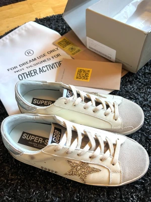 Golden Goose sneakers med glitterstjärna - Golden Goose sneakers i vitt skinn med beige mockadetaljer och silverfärgad häl. Sidan pryds av en glittrig guldstjärna som ger extra edge. Klassisk snörning och platt sula. Perfekta för dig som vill sticka ut med stilrena men ändå unika skor. Helt nya och oanvända.