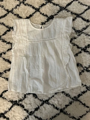 Vit blus med volang från Kappahl - Säljer en söt vit blus från Kappahl i storlek 158. Blusen har volangdetaljer längs axlarna och fina broderade sömmar framtill. Den är gjord i ett lätt och luftigt bomullstyg, perfekt för varma dagar. Rund halsringning och ärmlös design ger en fräsch och stilren look.