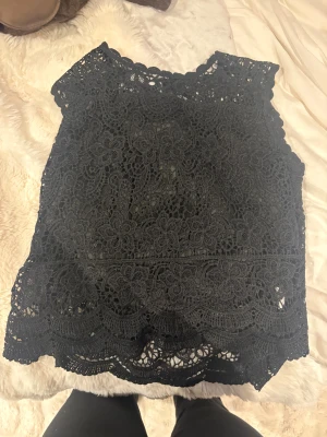 Svart spetsblus från Miss Girld L/XL - Supersnygg svart blus från Miss Girld i storlek L/XL. Blusen är helt täckt av detaljerad spets med blommigt mönster och har en scallop-kant längst ner. Den är ärmlös och har en rundad halsringning. Perfekt för dig som gillar statement-plagg med mycket detaljer.
