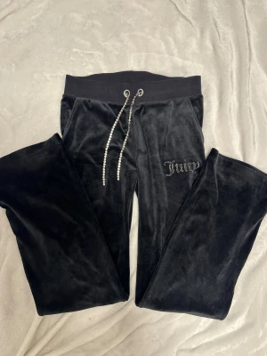 Svarta velourbyxor Juicy Couture S - Svarta mjukisbyxor från Juicy Couture i storlek S. Byxorna är i mjuk velour med glittrande strass-snörning i midjan och Juicy-logga i strass på benet. De har normal passform och bred resår i midjan för extra komfort.