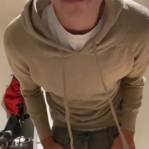 Beige hoodie med snörning - Säljer en stilren beige hoodie med huva som är lång ärmad. Tröjan har snörning vid halsen och är tillverkad i mjukt stickat material. Passformen är normal och den är perfekt för chill dagar. Enkel design utan tryck eller mönster. Rekommenderar för dig som är runt 170.