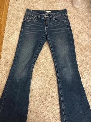 Blå bootcut jeans med broderade fickor - Snygga blå jeans från Gina med bootcut passform och låg midja. Jeansen har detaljerade broderier och dekorativa knappar på bakfickorna, samt kontrastsömmar. 