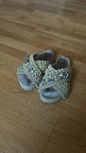 Inuikii tofflor - Unika beige sandaler med breda, grovt stickade band och stora glittrande pärlor i olika former. Sulan är platt och öppen tå ger en chill vibe. Perfekt för dig som gillar boho-stil och vill sticka ut med coola detaljer.