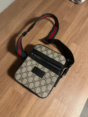 Gucci axelväska med GG-mönster - Snygg axelväska från Gucci med klassiskt GG-mönster i beige och brunt. Väskan har svarta detaljer, dragkedja framtill och en bred axelrem i grönt och rött. Perfekt storlek för det viktigaste och riktigt trendig design.