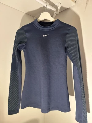 Nike Pro blå långärmad träningströja - Nike Pro Therma-Fit långärmad träningströja i mörkblått med diskret randiga detaljer på ärmarna och vit Nike-logga på bröstet. Slim passform och tillverkad i polyester för att hålla dig varm under träningen. Perfekt för dig som gillar sportig stil.