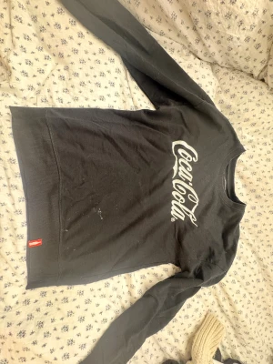 Svart Coca-Cola sweatshirt i bomull - Svart sweatshirt med klassisk Coca-Cola-logga i vitt på bröstet. Tröjan har långa ärmar med ribbade muddar och en liten röd detalj vid nederkanten. Tillverkad i mjuk bomull, perfekt för chill dagar. Rund hals och relaxed passform.