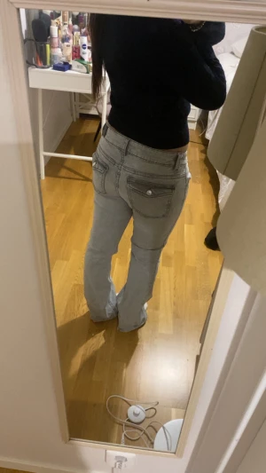 Ljusgrå bootcut jeans med fickdetaljer - Säljer ett par ljusgrå jeans med bootcut passform och låg midja. Jeansen har snygga fickdetaljer med knapp på bakfickorna och klassisk femficksdesign. Materialet är jeans och de har en stilren look som passar till många outfits.