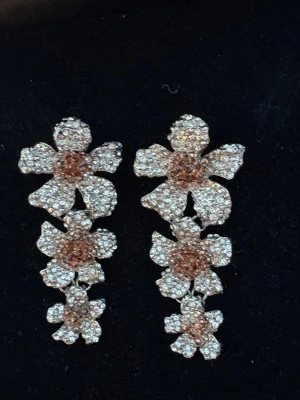 Glittriga blomformade örhängen i silver - Säljer ett par långa örhängen i form av tre sammankopplade blommor. Örhängena är täckta av små glittrande stenar i silver och roséguld, vilket ger en riktigt lyxig känsla. Perfekta för dig som vill sticka ut med ett unikt och blommigt smycke.