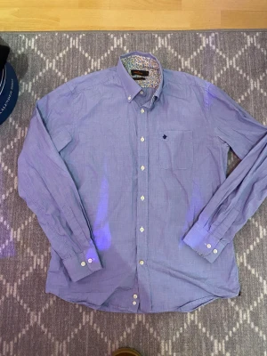 Blå skjorta från Morris med blommigt foder - Snygg blå skjorta från Morris med diskreta vita ränder och blommigt foder i kragen. Klassisk button-down-krage, bröstficka med broderad logga och vita knappar. Tillverkad i bomull, perfekt för dig som gillar stilrena detaljer.