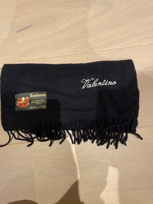 Svart halsduk från Valentino i ull - Stilren svart halsduk från Valentino med broderad logga och klassiska fransar i kanterna. Gjord i mjuk lammull som känns lyxig och varm. Perfekt att slänga över axlarna när det är kallt ute. Snygg och enkel design som funkar till de flesta jackor.